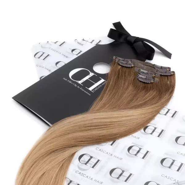 Cascata Hair Extensions - Honey Melt Brownie Full Set - Image of blonde hair extensions sitting on Cascata branded wrapping paper which is white with black Cascata logos repeated across it